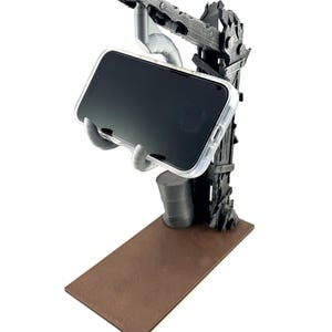 以下が含まれることがあります： グレーの3Dプリント製スマートフォンスタンド。ポスト・アポカリプス風のクレーンデザインで、質感のある垂直サポート、土台の小さな樽、そして透明なケースに入ったスマートフォンを保持する手形のクレードルが、茶色のベースの上に配置されています。