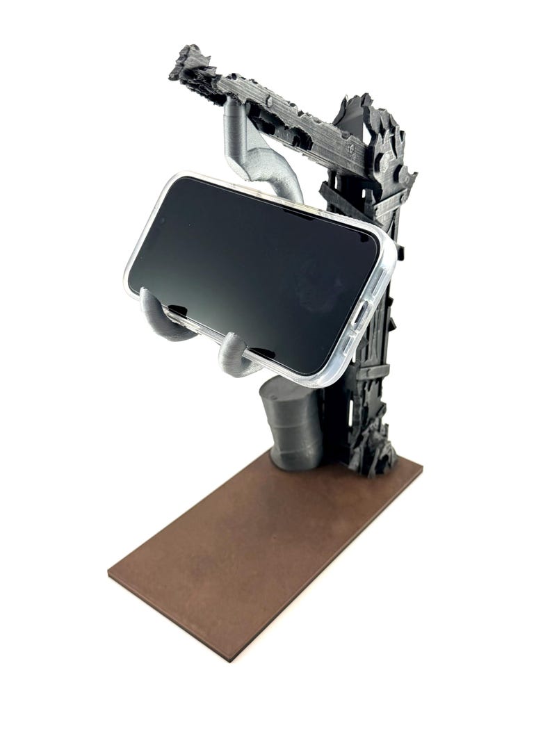 以下が含まれることがあります： グレーの3Dプリント製スマートフォンスタンド。ポスト・アポカリプス風のクレーンデザインで、質感のある垂直サポート、土台の小さな樽、そして透明なケースに入ったスマートフォンを保持する手形のクレードルが、茶色のベースの上に配置されています。