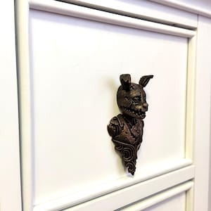 Pode incluir: Puxador de armário decorativo em tom bronze, apresentando uma cabeça de personagem estilizada e perturbadora com orelhas de coelho e um sorriso esquelético. O puxador é montado em uma porta de armário branca, adicionando uma estética gótica única.