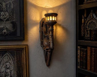 May include: A bronze-colored wall sconce with a skull design and a torch-like light fixture. The sconce is mounted on a textured wall, casting a warm glow. The design incorporates intricate details and a gothic aesthetic.