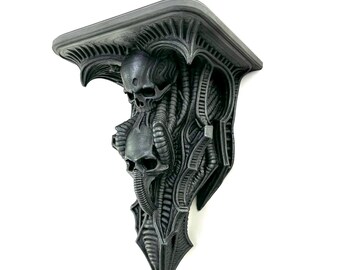 May include: A dark gray, wall-mounted shelf with a gothic, biomechanical design. The shelf features three skulls and intricate, organic-looking details. The shelf is made of a solid material and is mounted on a white wall.