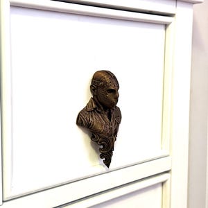 Pode incluir: Um busto de cor bronze de uma figura usando uma máscara de hóquei, uma camisa abotoada e um casaco. O busto é montado em uma superfície branca. O busto tem uma base decorativa e ornamentada.
