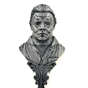 May include: A monochromatic gray wall-mounted bust depicting a figure wearing a mask with deep-set eyes and textured hair. The figure wears a collared shirt, and the base features an ornate, symmetrical scrollwork design with intricate carvings.