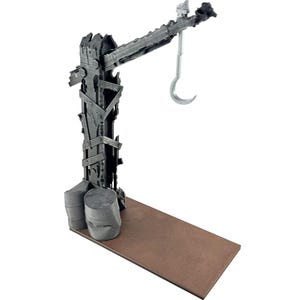 May include: A dark gray, weathered crane structure featuring a vertical post reinforced with metal straps and two cylindrical barrels at the base. A horizontal arm extends from the top, supporting a silver-colored metal hook hanging downward.