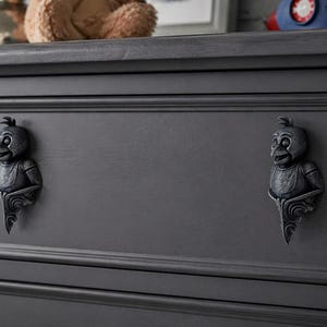 Op de afbeelding: Een donkergrijze commode met twee lades, elk versierd met een decoratieve vogelvormige handgreep. De handgrepen hebben een vintage ontwerp. De commode maakt deel uit van een kinderkamerset.