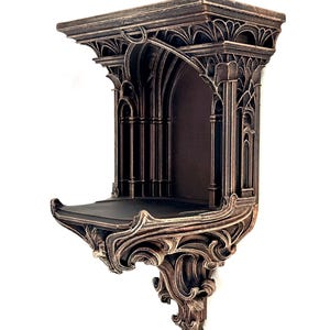 May include: A wall-mounted shelf featuring a dark bronze finish with intricate architectural details, including arched columns and ornate scrollwork supports. The structure has a flat top surface and a recessed central alcove area.