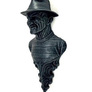 Pode incluir: Busto cinza escuro de um personagem usando um chapéu fedora e um suéter listrado. O busto tem uma superfície detalhada e texturizada e é montado em uma base decorativa e ornamentada. Escultura com tema de terror.