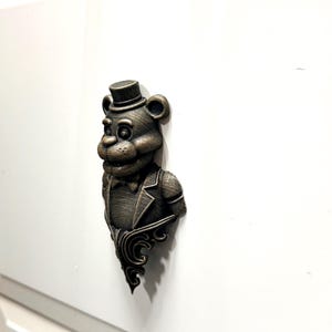 Pode incluir: Puxador de armário em bronze em forma de urso com cartola e gravata borboleta. O urso está usando um paletó e tem uma base ornamentada. O puxador está montado em uma porta de armário branca.