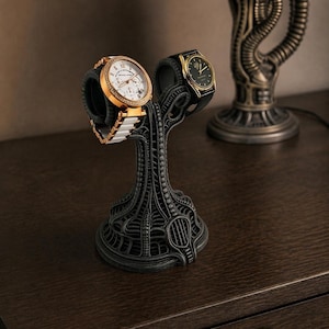 Puede incluir: Un soporte negro para relojes con un diseño orgánico intrincado sostiene dos relojes. Uno tiene un bisel dorado y una correa blanca, el otro una correa negra y detalles dorados. El soporte está sobre una superficie de madera oscura.