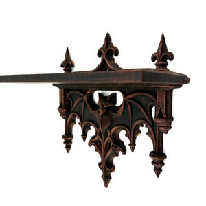 May include: A wall-mounted shelf featuring a dark brown finish with black highlights. The support structure displays a central bat figure with outstretched wings, surrounded by intricate pointed arches and three decorative finials on top.