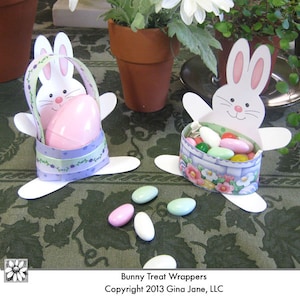 Op de afbeelding: Twee witte, konijnvormige traktatiehouders met bloemmotieven. De ene houder is gevuld met roze snoepjes en de andere met pastelkleurige snoepjes. De tekst "Bunny Treat Wrappers Copyright 2013 Gina Jane, LLC" staat onderaan de afbeelding.