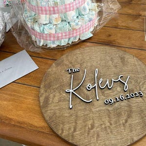 May include: A circular wooden sign with the inscription "The Kofews" and the date "09.16.2023" in white. A diaper cake and cards are in the background. The sign is made of wood with a natural grain.