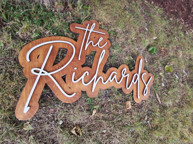Pode incluir: Uma placa de madeira com as palavras "The Richards" em letras brancas em cursiva. A placa &eacute; marrom e tem um visual r&uacute;stico.