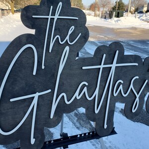Pode incluir: Uma placa de madeira preta com letras brancas que diz "The Ghattasi's". A placa est&aacute; apoiada em um suporte de metal preto.