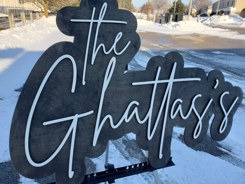 Pode incluir: Uma placa de madeira preta com letras brancas que diz "The Ghattasi's". A placa est&aacute; apoiada em um suporte de metal preto.