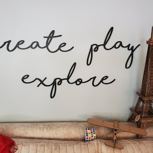 May include: Black wooden wall decor with the words "create play explore" in a cursive font. A wooden model of the Eiffel Tower and a wooden airplane are on a tan couch.