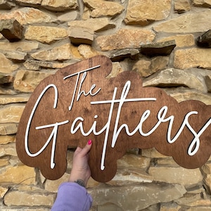 Pode incluir: Uma placa de madeira com fundo marrom e letras brancas. A placa diz "The Gaithers" em uma fonte cursiva.