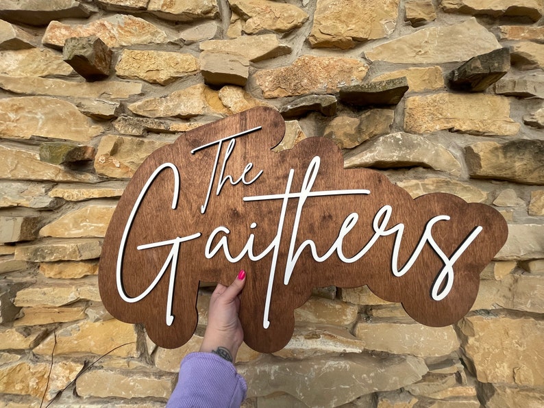 Pode incluir: Uma placa de madeira com fundo marrom e letras brancas. A placa diz "The Gaithers" em uma fonte cursiva.