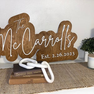 Pode incluir: Uma placa de madeira com o texto "The Mc Carrolls Est. 2.26.2023" em letras brancas. A placa tem a forma de um nome de fam&iacute;lia estilizado.