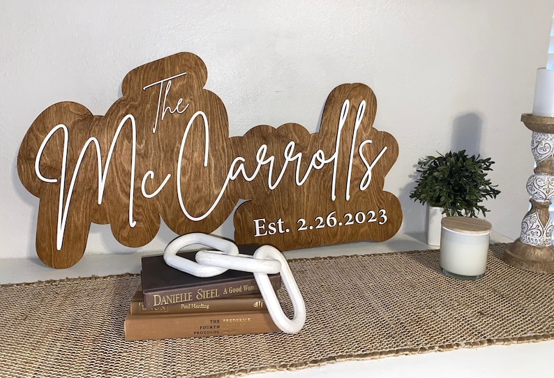 Pode incluir: Uma placa de madeira com o texto "The Mc Carrolls Est. 2.26.2023" em letras brancas. A placa tem a forma de um nome de fam&iacute;lia estilizado.