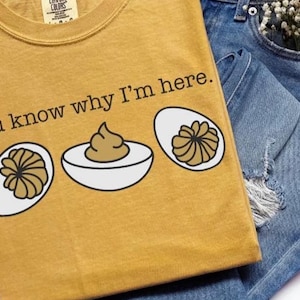 May include: Mustard yellow t-shirt with the text "You know why I'm here" and a graphic of deviled eggs. The shirt is paired with blue denim shorts. The shirt has a crew neck and short sleeves.