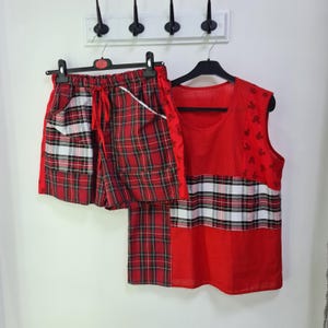 May include: A red plaid pyjama set with a matching sleeveless top and shorts. The shorts have a drawstring waist and pockets. The top features a polka dot pattern and Mickey Mouse silhouettes. The set is hanging on a white wall.