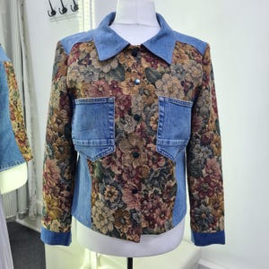 Könnte beinhalten: Jeans- und Blumenserie-Jacke. Die Jacke hat einen hellblauen Jeanskragen, Schultern und Taschen mit einem floralen Wandteppichdesign auf der Vorderseite und den Ärmeln. Das Blumenmuster umfasst Brauntöne, Rot und Grün.