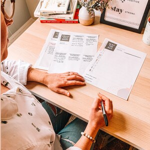 May include: A person is sitting at a desk with a planner and a pen. The planner has the words "Slay the Day" on the cover. The person is writing in the planner. There are books and a plant on the desk. The wall behind the person has a framed sign that says "stay focused, stay positive, stay strong."