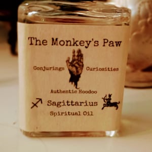 May include: A small glass bottle with a brown label that reads "The Monkey's Paw Conjuring Curiosities Authentic Hoodoo Sagittarius Spiritual Oil". The label also features a hand with three fingers extended and a small image of a centaur with a bow and arrow.