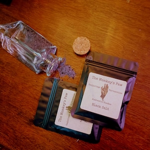 May include: A clear glass bottle with a cork stopper, containing dark purple salt, alongside two black resealable pouches. Each pouch features a white label with the text The Monkey's Paw, Conjuring Curiosities, Authentic Hoodoo, Black Salt.