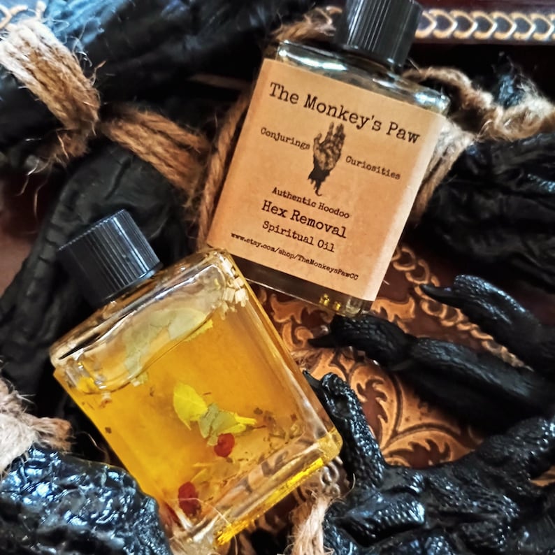 May include: Two small glass bottles with cork stoppers and gold liquid inside. The bottles are labeled "The Monkey's Paw Conjurings Curiosities Authentic Hoodoo Hex Removal Spiritual Oil www.etsy.com/shop/TheMonkeysPawCO"