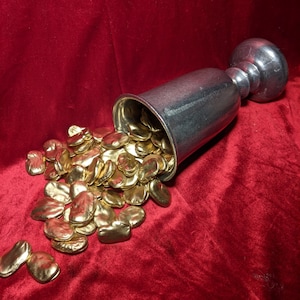 May include: A silver goblet spills out gold-colored beans onto a red velvet surface.
