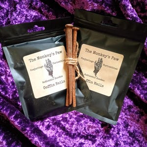 May include: Two black pouches with the text "The Monkey's Paw" and "Coffin Nails" on a purple velvet surface. A bundle of rusty nails is tied with twine between the pouches. The pouches also have a graphic of a hand.