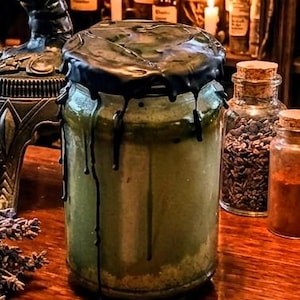 May include: A clear glass jar filled with a pale green substance, sealed with a thick layer of black wax that drips down the sides of the container. The jar has a cylindrical shape and contains a textured, light-colored sediment at the bottom.