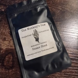 Puede incluir: Una bolsa negra con una etiqueta blanca que dice "The Monkey's Paw Conjuring Curiosities Authentic Hoodoo Snake Shed www.etsy.com/shop/TheMonkeysPawCo"