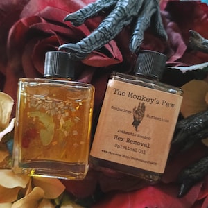 May include: Two small glass bottles with gold liquid and black caps. The bottles are labeled "The Monkey's Paw Conjuring Curiosities Authentic Hoodoo Hex Removal Spiritual Oil www.etsy.com/shop/TheMonkeysPaw".