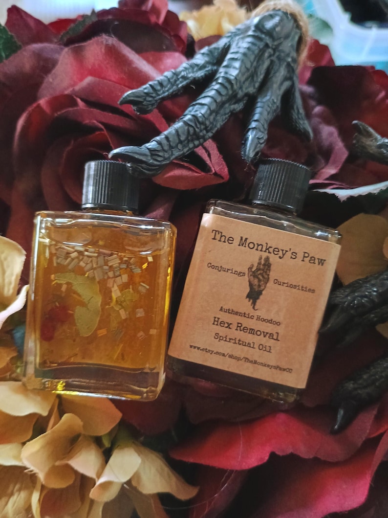 May include: Two small glass bottles with gold liquid and black caps. The bottles are labeled "The Monkey's Paw Conjuring Curiosities Authentic Hoodoo Hex Removal Spiritual Oil www.etsy.com/shop/TheMonkeysPaw".