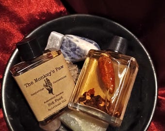 Könnte beinhalten: Zwei kleine Glasflaschen gefüllt mit einer goldenen Flüssigkeit und verschiedenen Kräutern und Gewürzen. Die Flaschen sind mit "The Monkey's Paw Conjurings Curiosities Authentic Hoodoo Hot Foot Spiritual Oil" beschriftet. Die Flaschen stehen in einer schwarzen Schale mit ein paar kleinen Steinen.