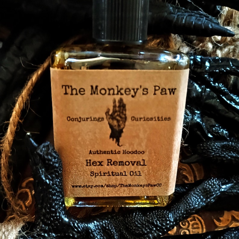 May include: A small brown bottle with a label that reads "The Monkey's Paw Conjuring Curiosities Authentic Hoodoo Hex Removal Spiritual Oil www.etsy.com/shop/TheMonkeysPawCo". The bottle is sitting on a black surface.