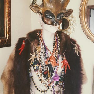 Puede incluir: Un maniquí con un abrigo de piel marrón y una variedad de collares, incluyendo cuentas, dijes y una máscara con cuernos. La máscara es marrón y tiene un aro de nariz dorado.