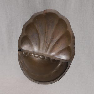 May include: A vintage, copper-toned metal mold in a seashell design. The mold has a scalloped top and a rounded bottom with a decorative beaded edge. The metal has a patina, showing signs of age and use.