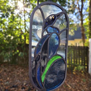 May include: Stained glass bird suncatcher with a dark metal frame. The bird features shades of blue, black, and green glass, with a yellow eye. The suncatcher is hanging from a blue string.