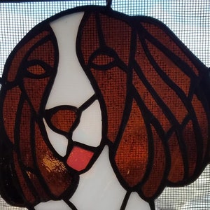 May include: Stained glass artwork depicting a dog's face. The dog has brown ears and a white face with a red tongue. The artwork is framed with black lines and set against a mesh background.
