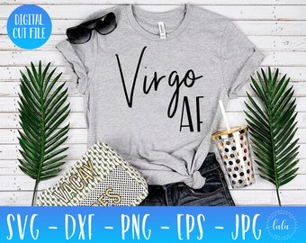 May include: A gray t-shirt with the text "Virgo AF" in black script font. The shirt is laying on a white wooden surface with a pair of sunglasses, a clutch purse, and a clear tumbler with gold polka dots.