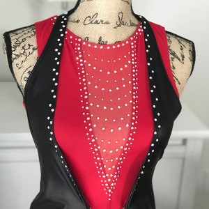 May include: A red sleeveless leotard with a black vest. The leotard has a sheer panel in the front with rhinestones. The vest is open in the front and has rhinestones along the edges.