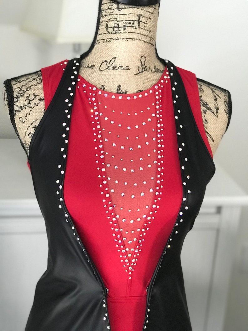 May include: A red sleeveless leotard with a black vest. The leotard has a sheer panel in the front with rhinestones. The vest is open in the front and has rhinestones along the edges.