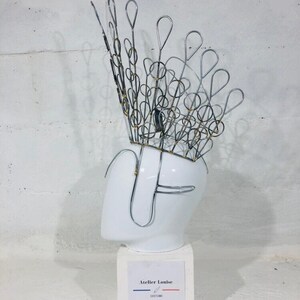 May include: A silver wire crown with a peacock design, displayed on a white mannequin head. The crown is adorned with small gold beads. The base of the mannequin is white and has a label that reads "Atelier Louise Costumes".