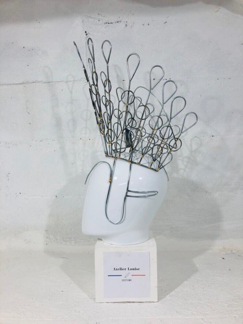 May include: A silver wire crown with a peacock design, displayed on a white mannequin head. The crown is adorned with small gold beads. The base of the mannequin is white and has a label that reads "Atelier Louise Costumes".