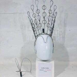 May include: A silver wire crown with gold accents on a white mannequin head. The crown is on a white pedestal with the text "Atelier Louise Costume" printed on a label.
