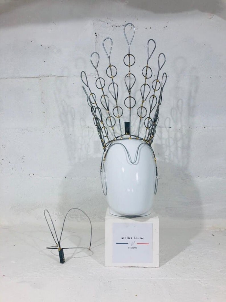 May include: A silver wire crown with gold accents on a white mannequin head. The crown is on a white pedestal with the text "Atelier Louise Costume" printed on a label.